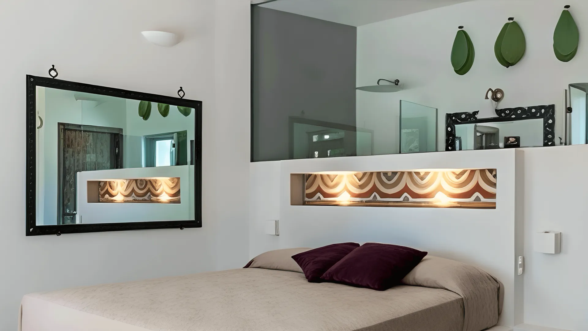 Modern bedroom designed for family luxury accommodation Salento, featuring a beige bed, purple pillows, built-in headboard niche with decor and spotlights, black-framed mirror, white walls, glass partition, and three green leaf-shaped wall decorations.