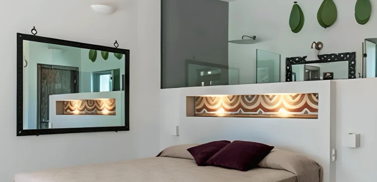 Modern bedroom designed for family luxury accommodation Salento, featuring a beige bed, purple pillows, built-in headboard niche with decor and spotlights, black-framed mirror, white walls, glass partition, and three green leaf-shaped wall decorations.
