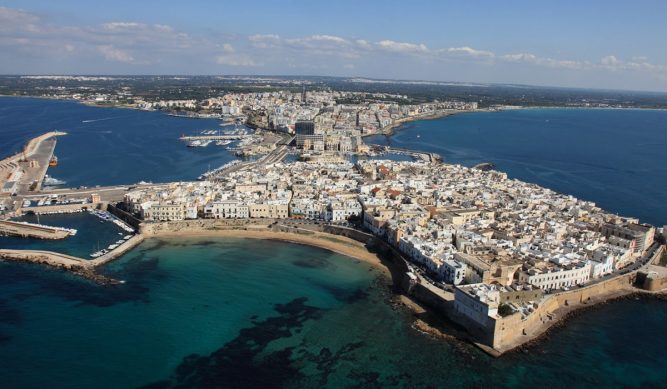 Aerial view of a coastal Italian town with dense white buildings, surrounded by turquoise sea. Near the old town’s ancient stone walls, a Gallipoli luxury villa exterior stands out, overlooking the marina and modern city under a clear blue sky.