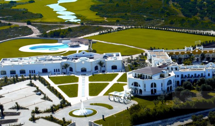 Aerial view of the luxurious white Acaya Golf Resort & Spa featuring manicured gardens, walking paths, a unique-shaped pool, and green lawns—perfect for golf in Salento—surrounded by natural landscape, trees, fields, and a winding waterway.