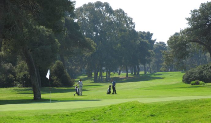 Two golfers stand on a lush green fairway at Golf in Salento Riva dei Tassali, their bags nearby under tall trees. Sunlight streams through the branches, casting gentle shadows and creating a peaceful golf course scene near the white-flagged hole.