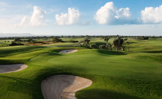 A lush green golf course under a blue sky with fluffy white clouds. Experience golf in Salento at San Domenico Golf, featuring sand bunkers, a manicured putting green with flagstick, and gently rolling hills with scattered trees.