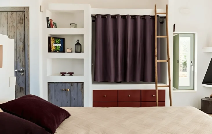 A cozy bedroom, inspired by the best luxury villa Salento, features a beige bed with dark purple pillows, built-in white shelving with books and art, red drawers, a wooden ladder, rustic cabinet doors, and purple curtains for a modern eclectic look.
