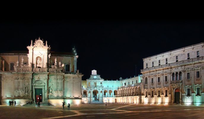 A grand, ornately decorated cathedral stands illuminated at night in a spacious, empty plaza in Lecce. Historic buildings with arched windows and columns flank the scene. Soft lighting highlights details as a few figures walk near the cathedral entrance.