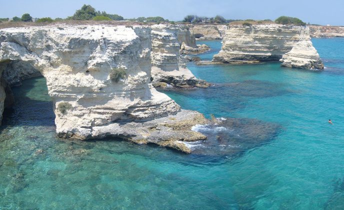 White limestone cliffs near Manduria rise above clear turquoise water along a rocky coastline. The sea is calm, revealing submerged rocks beneath the surface. Sparse vegetation grows atop the cliffs under a clear blue sky.