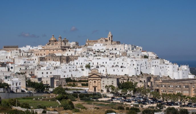 The hilltop town of Ostuni, with its tightly clustered white buildings, churches, and domes, stands under a blue sky. Green trees and parked cars lie below, while the sea is faintly visible on the horizon, enhancing its Mediterranean charm.