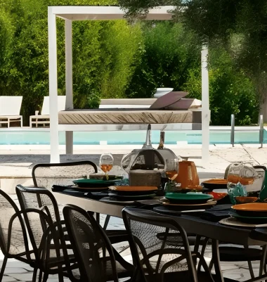 A stylish outdoor dining table is set with colorful plates and glasses under a tree at an exclusive holiday villa in Salento. Black chairs surround the table, with a swimming pool, white lounge beds, and lush greenery creating a relaxing atmosphere.