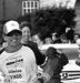 A male athlete in race gear smiles as he runs toward the finish line, wearing sunglasses, a cap, and bib 1986. Spectators on the left cheer and clap. The event feels energetic with banners, a parked car, and even a villa mia review sign in the background.