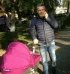 A man in a dark jacket and jeans walks in a sunlit park, pushing a bright pink baby stroller while talking on his cellphone—reminiscent of relaxing days mentioned in a Salento Villa Mia review, surrounded by trees and greenery.