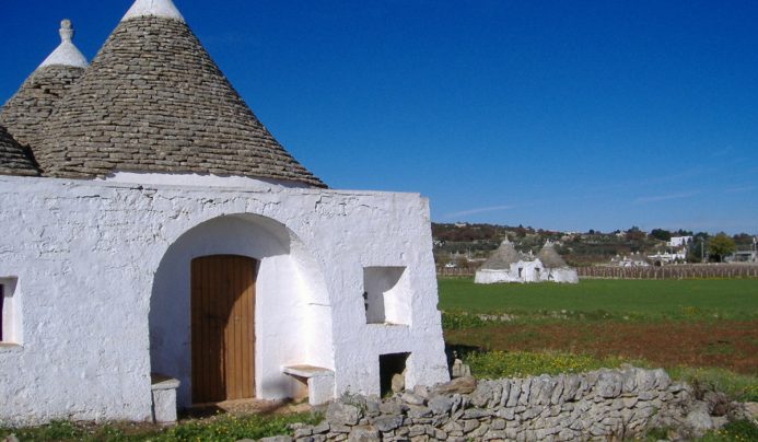 A white stone house with a conical gray stone roof stands in Valle d'Itria, fronting a green field under a blue sky. The house features an arched doorway, wooden door, and small windows. Similar cone-roofed buildings dot the background.
