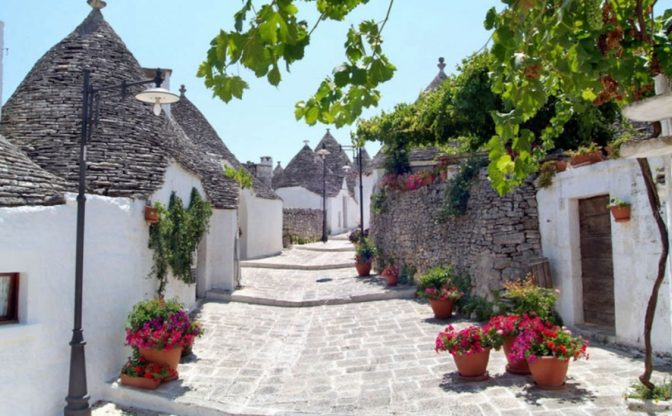 A narrow stone street in Valle d'Itria, Puglia, lined with whitewashed houses featuring conical stone roofs. Flower pots and green vines decorate the pathway, while a lamp post and leafy grapevine hang overhead beneath a clear blue sky.