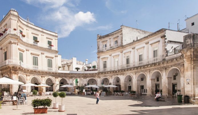 A sunlit Italian piazza in Martina Franca, Valle d’Itria, with curved white stone buildings featuring arched porticoes and balconies. Outdoor café tables dot the square as people stroll among potted trees under a brilliant blue sky.