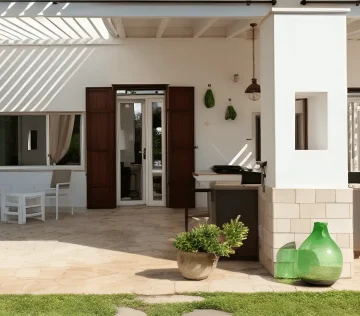 A modern outdoor patio at Vila Mia Salento Luxury features beige stone flooring, white walls, and brown wooden shutters. A dining table, potted plants, a green glass vase, and grill sit in sunlight with striped shadows and grassy borders.