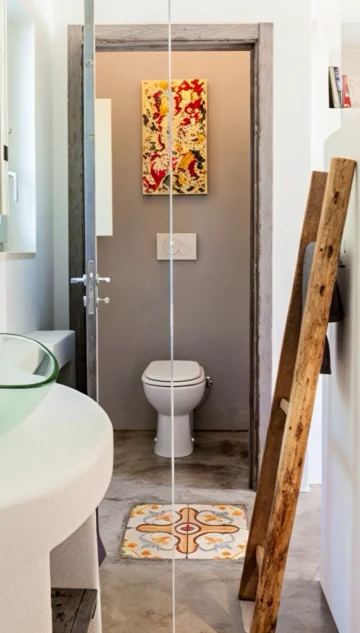Modern bathroom with a glass sink on a round counter, rustic wooden ladder leaning on a white partition, and a small room with a toilet. Colorful art and patterned tile decorate the toilet area. Light walls and gray floors create a bright, airy feel.