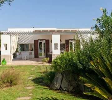 A luxury villa with pool in Salento, this modern white house with wooden shutters and a pergola-covered patio sits amid lush greenery. Elegant outdoor furniture, flowing white curtains, and a stone path complete the inviting garden retreat.