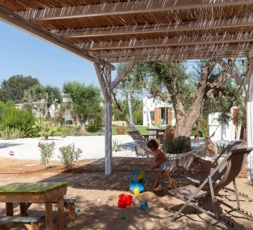 A child plays with colorful sand toys in a shaded outdoor sandbox beneath a pergola at a Puglia luxury seaside villa rental. The lush garden, hammock, and views of white buildings under the bright blue sky create an idyllic family retreat.
