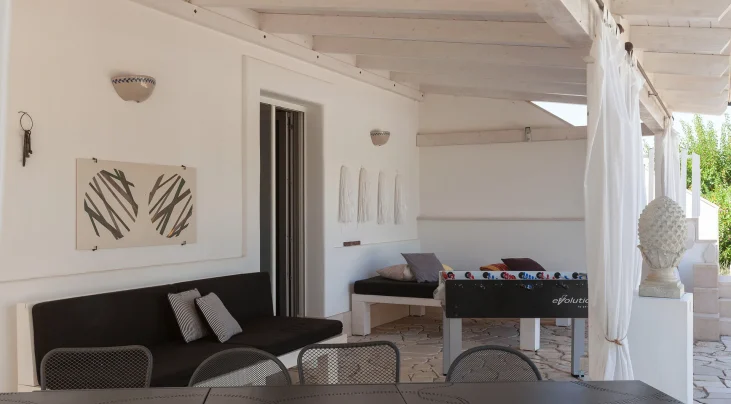 A modern covered patio at the best luxury villa Salento features white walls, wooden beams, a black sofa, decorative wall art, a foosball table, and a dining area with mesh chairs. Sheer curtains and stone flooring complete the relaxed, contemporary vibe.