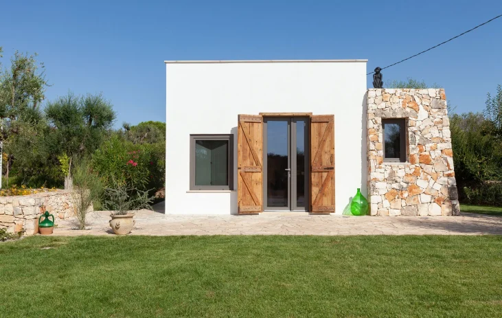 An exclusive holiday home in Salento, this small modern white house features wooden shutters and a unique stone section adorned with green glass bottles. Set on a stone patio amid lush grass and plants, it’s perfect under the clear blue sky.