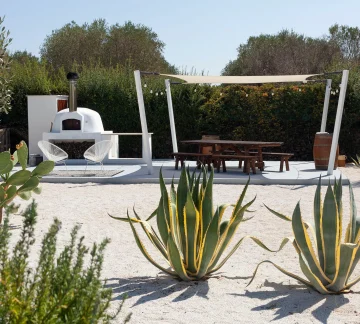 A modern outdoor patio features a white wood-fired pizza oven, picnic tables under a beige canopy, and tall agave plants—perfect to explore the magic of Salento from your private villa on a sunny day, surrounded by lush greenery.