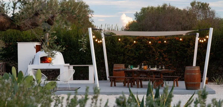 An outdoor patio perfect for Booking Your Dream Holiday Villa features a white pizza oven, a wooden dining table with benches, string lights overhead, and two barrels. Lush greenery and rustic touches create an inviting atmosphere for relaxation.
