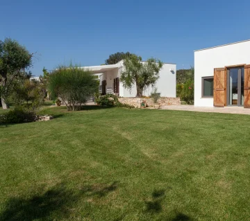 Modern white house with wooden doors and large windows, surrounded by a well-manicured green lawn. Trees, shrubs, and stone accents enhance the peaceful, spacious setting—ideal for a luxury villa with pool in Salento under a clear blue sky.