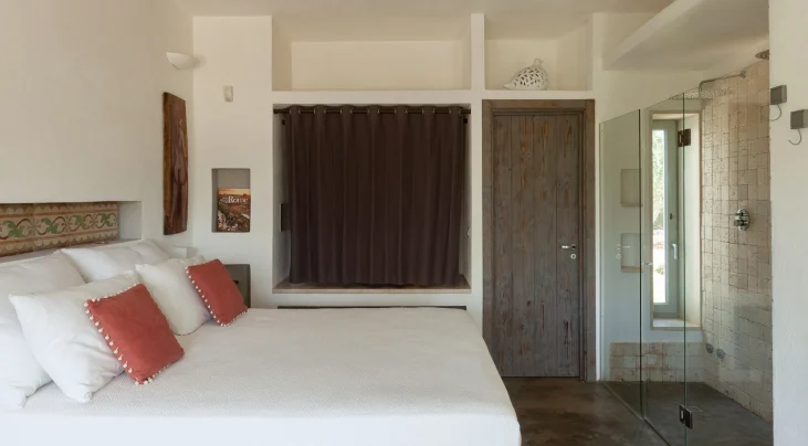 A minimalist bedroom in a luxury villa rental Salento coast, featuring a large bed with white linens and orange pillows. White walls, decorative tiles behind the headboard, and a glass-enclosed shower area complete this serene retreat.