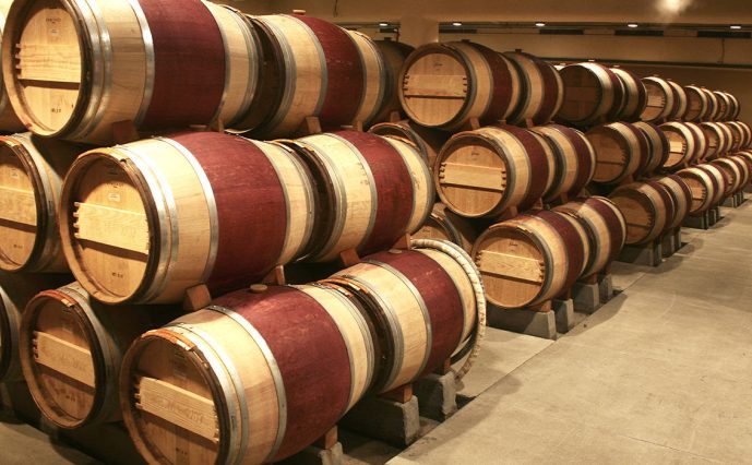 Rows of stacked wooden barrels, likely for aging vino-manduria Salento, are arranged on racks in a well-lit cellar with a concrete floor. The barrels show red stains from recent use, and the space is clean and organized.